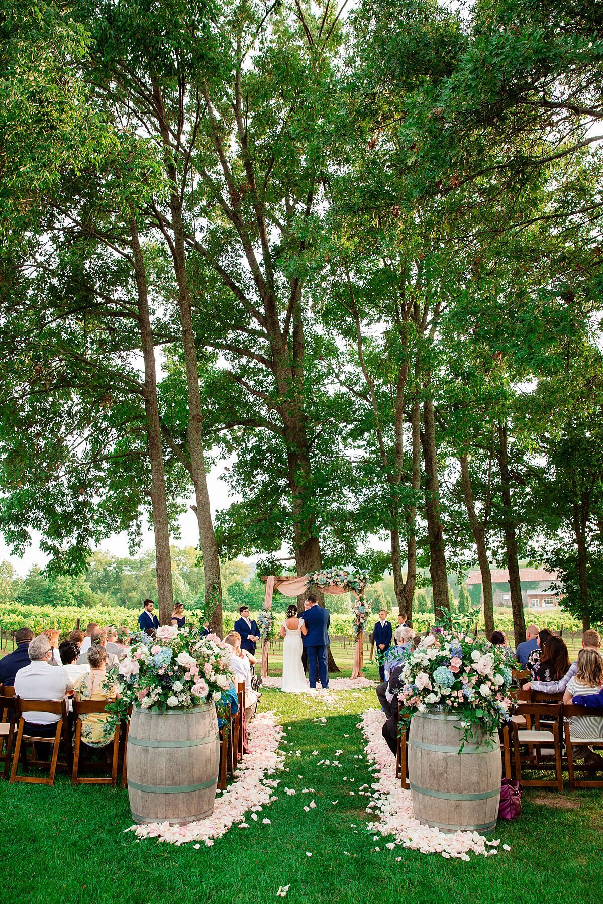 Wedding guests seated in brown fruit wood chairs watch the bride and groom exchange vows at their Arrington Vineyard wedding. Two wine barrels sit on either side of the aisle with large pink and blue floral arrangements. White ad blush rose petals line the center aisle and lead to a wooden arbor decorated with blush pink fabric and sprays of greenery, pink and blue flowers.