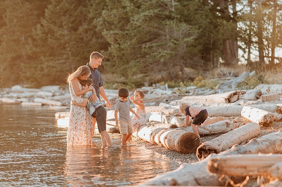 A happy family of 6 with 4 toddlers play in the water with driftwood all over the beach with their Vancouver Family Photographer