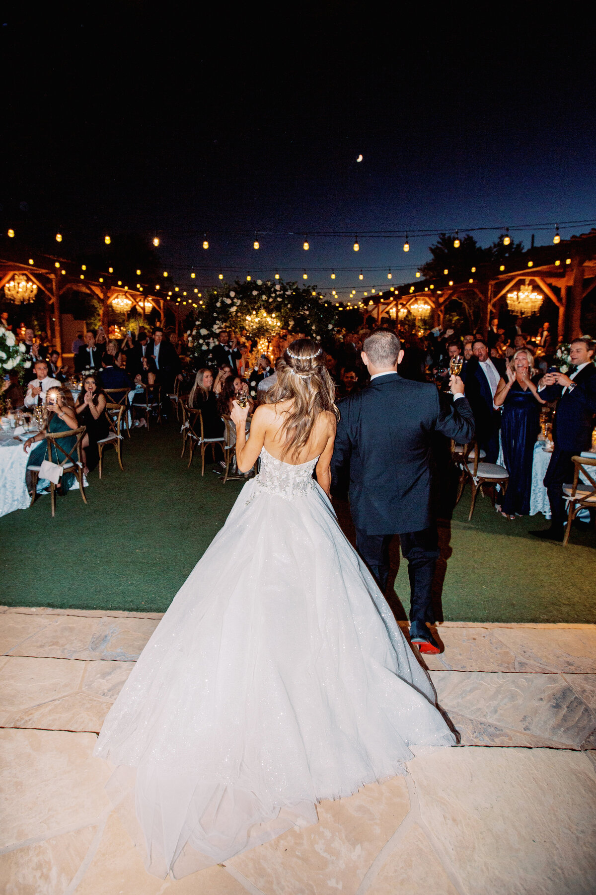 The bride and groom make a grand entrance under a canopy of twinkling string lights, setting the tone for a magical outdoor wedding reception.