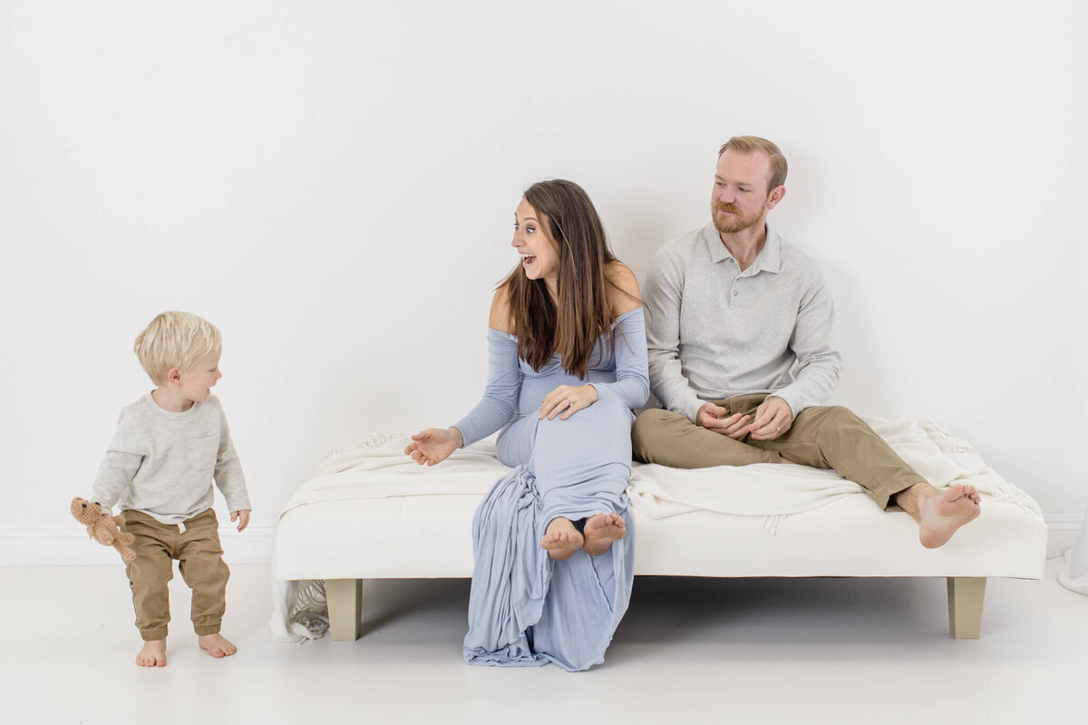 A woman in a light blue dress sits on a bed with a man in a gray sweater, both smiling at a toddler standing nearby, wearing a light gray shirt and brown pants. The scene is set in a bright, minimalistic room.
