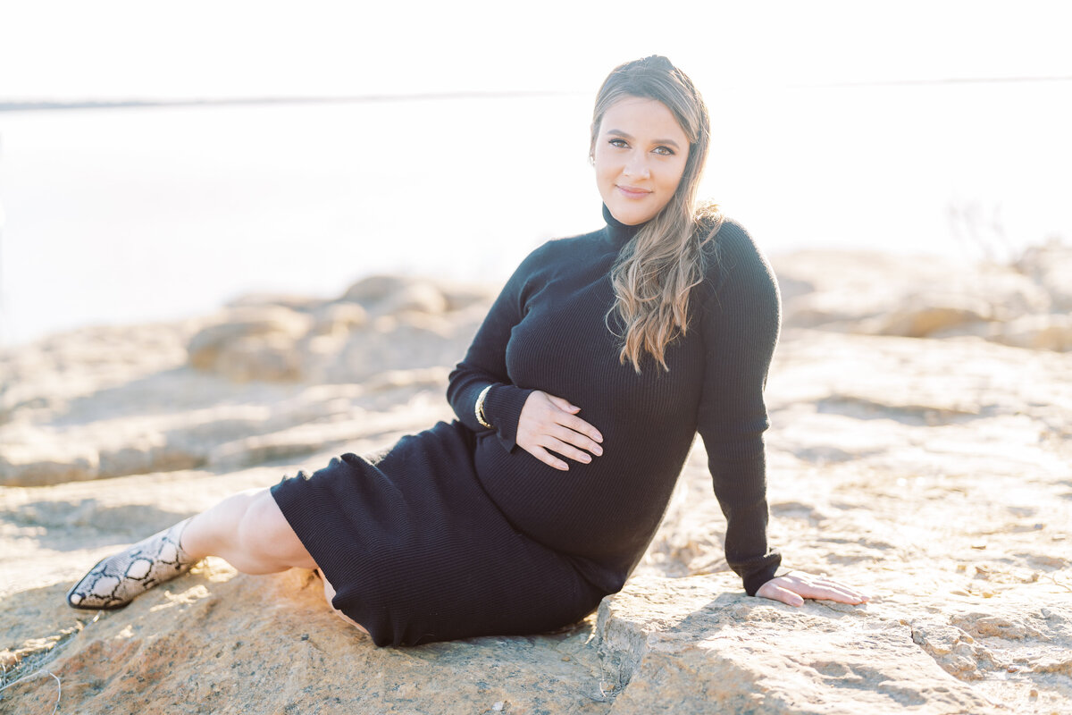 Portrait of a pregnant woman wearing black sitting on the sand with her hand over her belly at sundown.