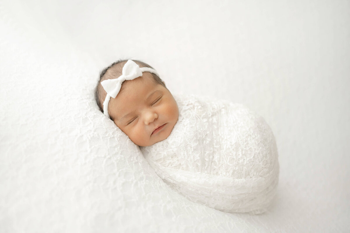 newborn baby girl wearing a white bow sleeping on a vintage lace blanket in an oklahoma city studio in the Wheeler District