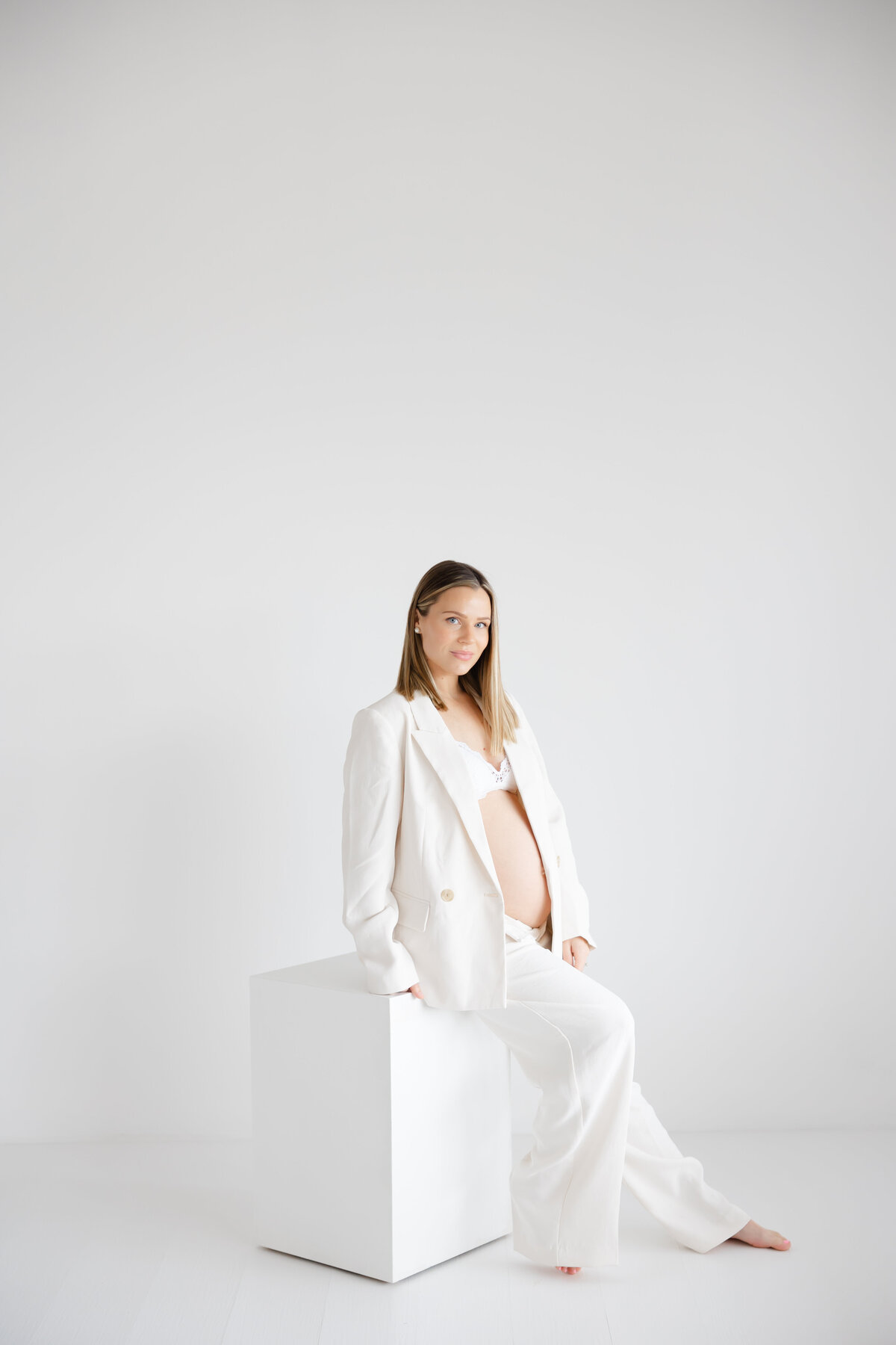 a pregnant mother sits on a white posing block in a white studio while wearing a white blazer and pantsuit matching set. She sits and glances at the camera with a serious face ad straight modern haircut.