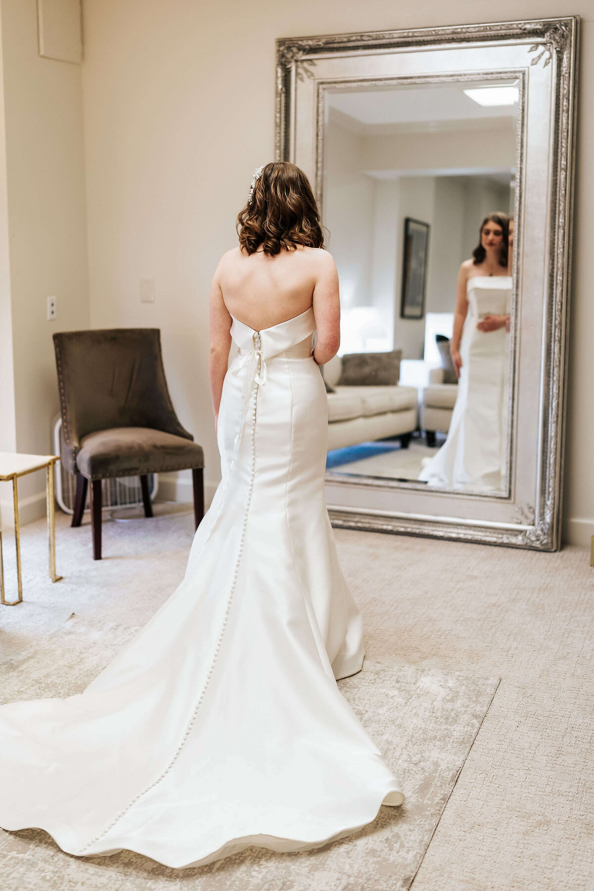 A bride in a strapless white wedding dress stands in front of a large mirror, viewing her reflection in a bright, elegantly furnished room.