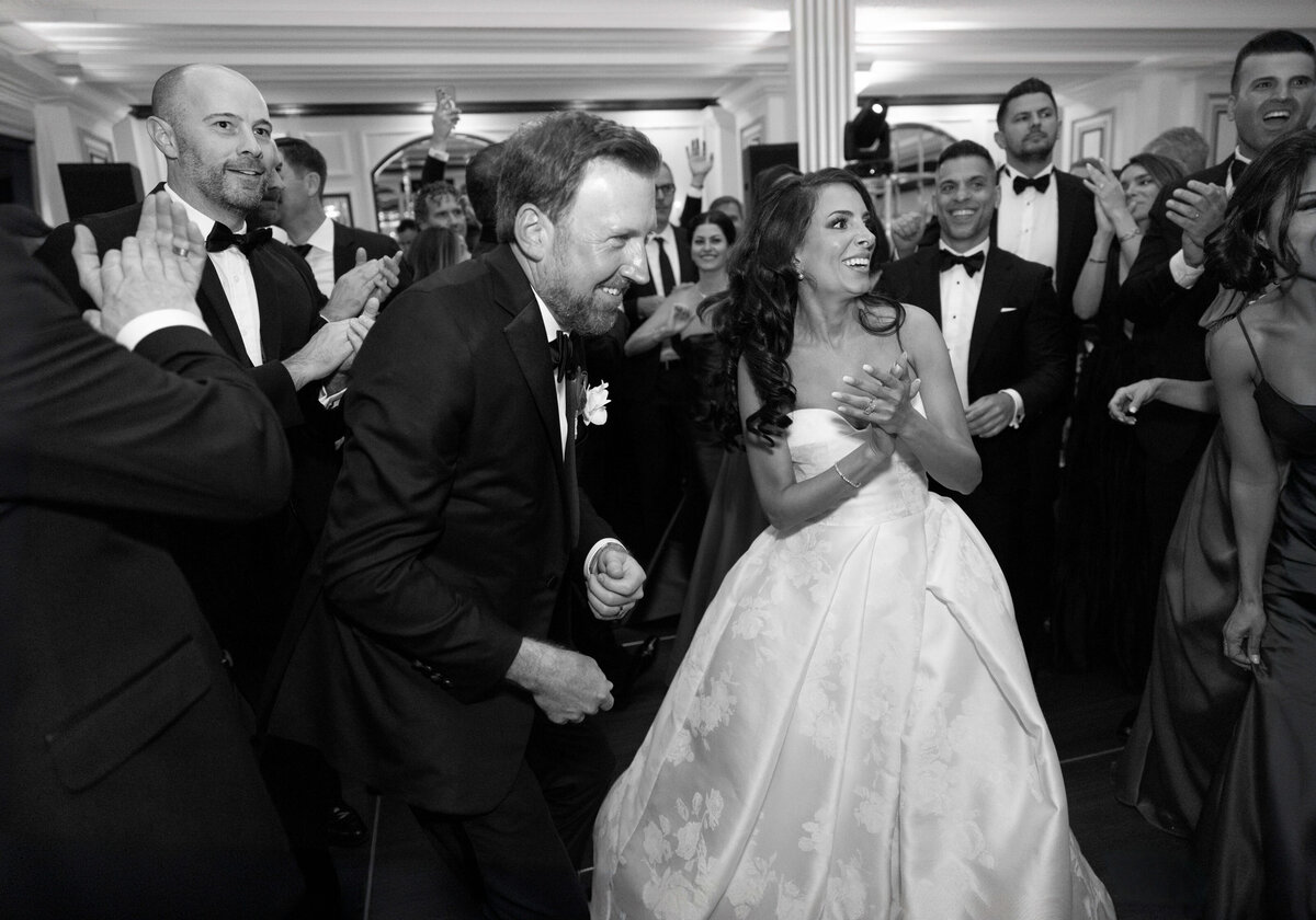 A bride in a flowing gown and a groom in a tuxedo dance joyfully, surrounded by applauding guests in formal attire at a wedding reception. The room is elegantly decorated, and everyone appears to be having a great time. Black and white photograph.