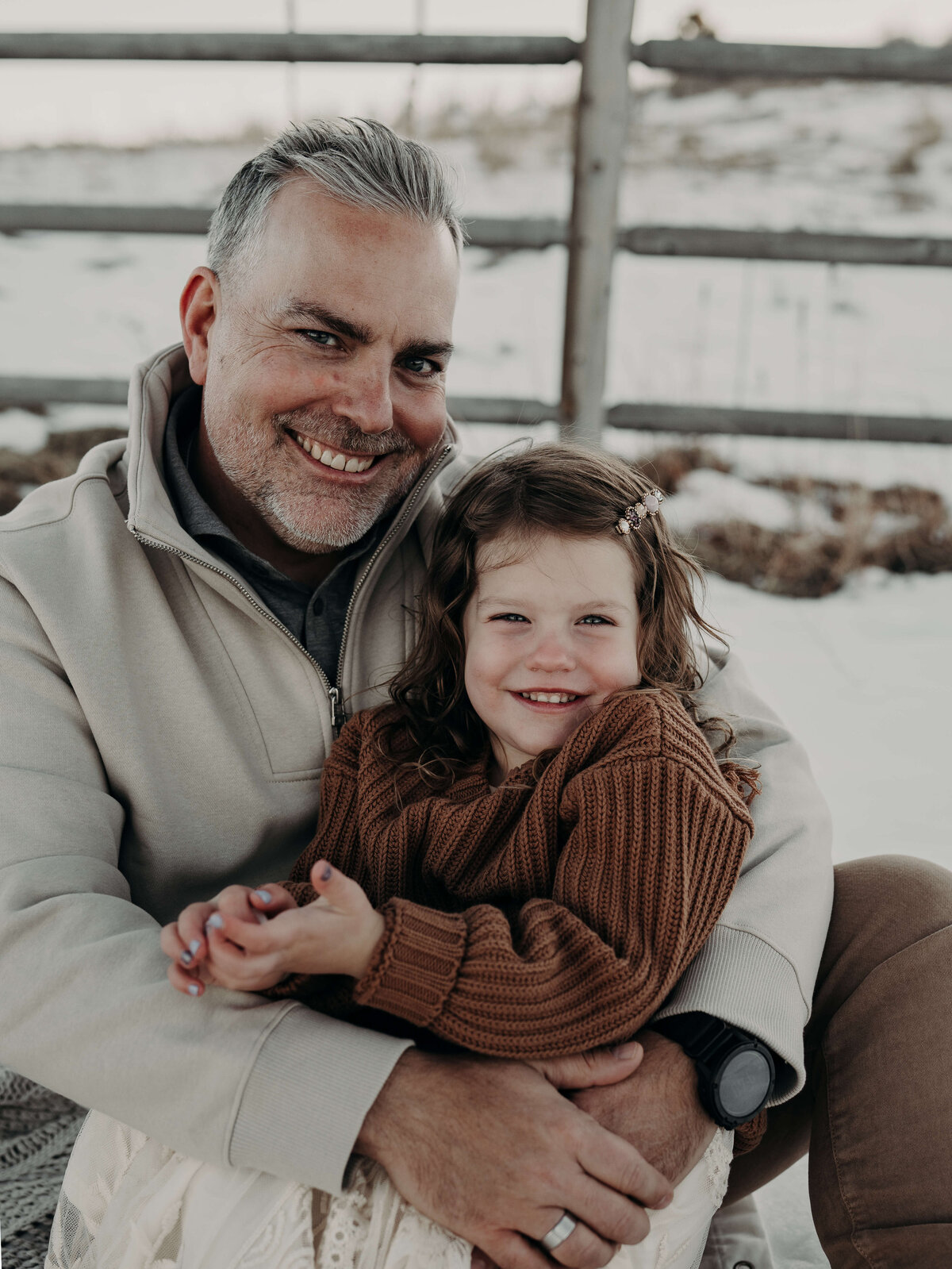 A middle-age dad holds his toddler daughter on his lap as they both smile towards the audience while she wears a giant brown fluffy sweater and he wears a cream color.