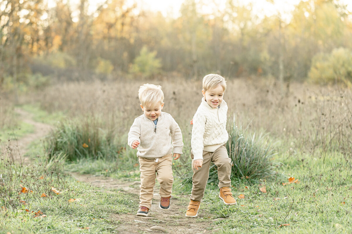 Two little boys dressed in khakis and cream sweaters running towards the camera in a field of tall grasses. Outdoor family photos in Oregon.