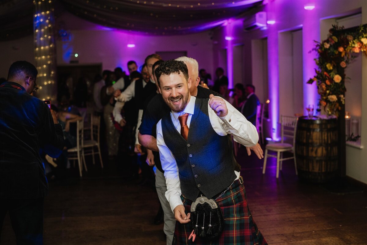 A large group of wedding guests dance in a line and walk through a large venue space. Fairy lights hang around them.