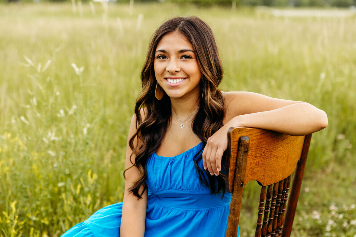 brunette teen girl smiling as she rest her arm on the back of a wooden chair while sitting in a pretty grassy field near Appleton WI