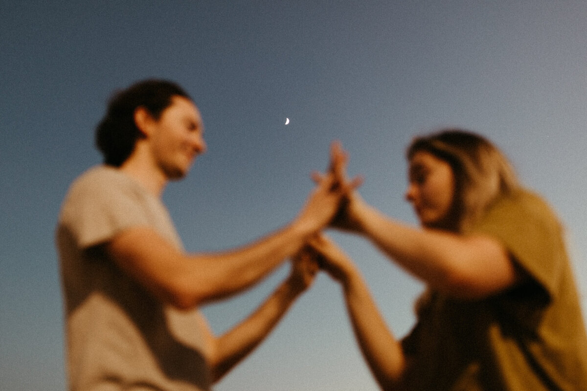 A guy and a girl are standing face to face and holding hands between them while the sky is getting dark behind them and the moon is shining.