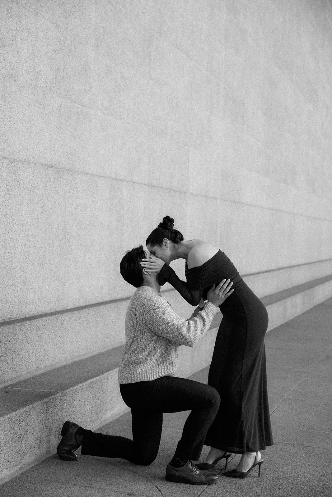 Man kneels down on one knee and proposes as she responds yes with a kiss. 