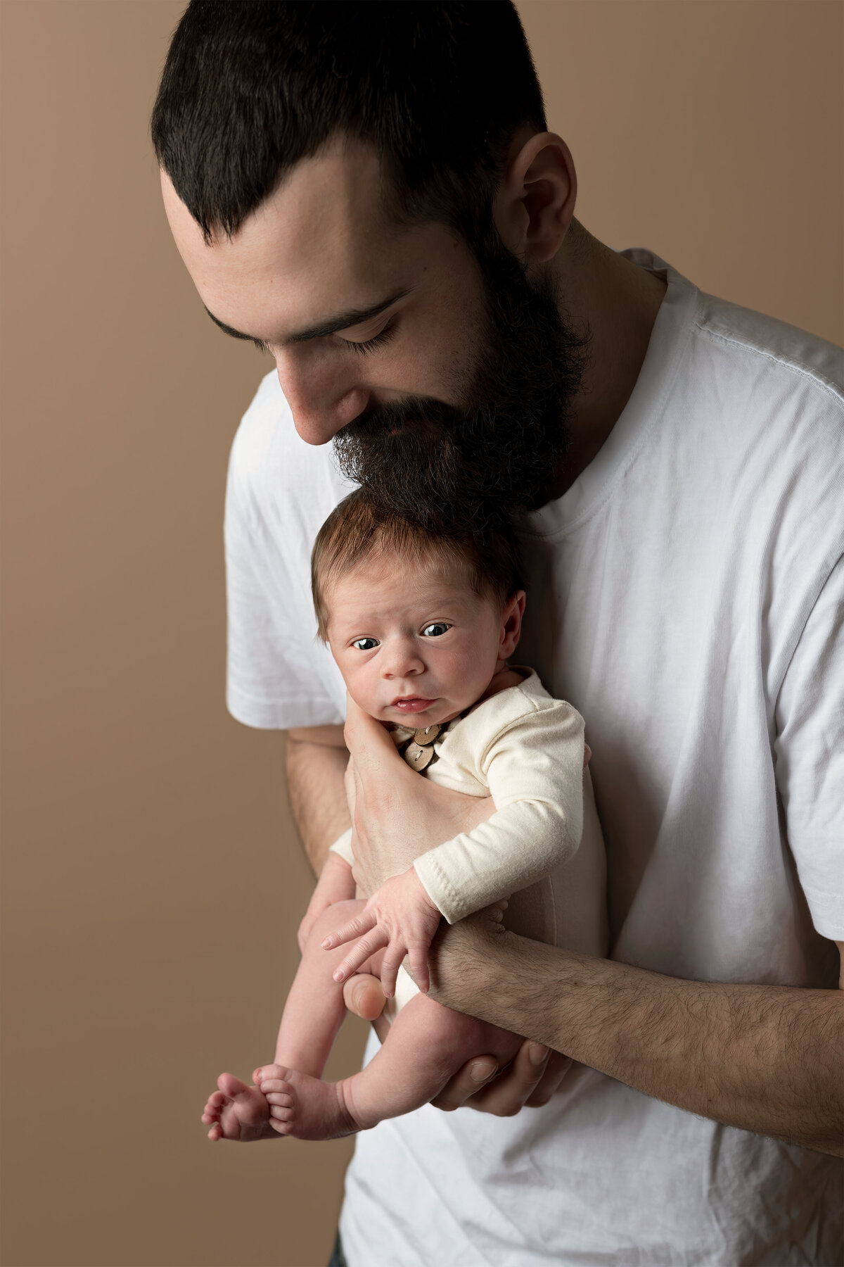 Emotive studio portrait of a bearded father tenderly holding his newborn baby in Paris, Ontario. Captured by Meraki Photographic Creations. Photographed in-studio at Meraki Photographic Creations in Paris, Ontario.