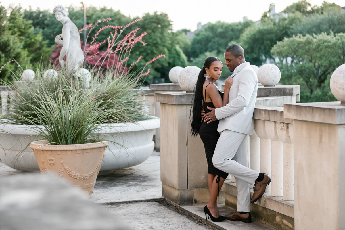 Engaged couple at Adriatica Village in Dallas during sunset, he leaning on a railing while she leans toward him looking down, surrounded by romantic European-inspired architecture and sculptures