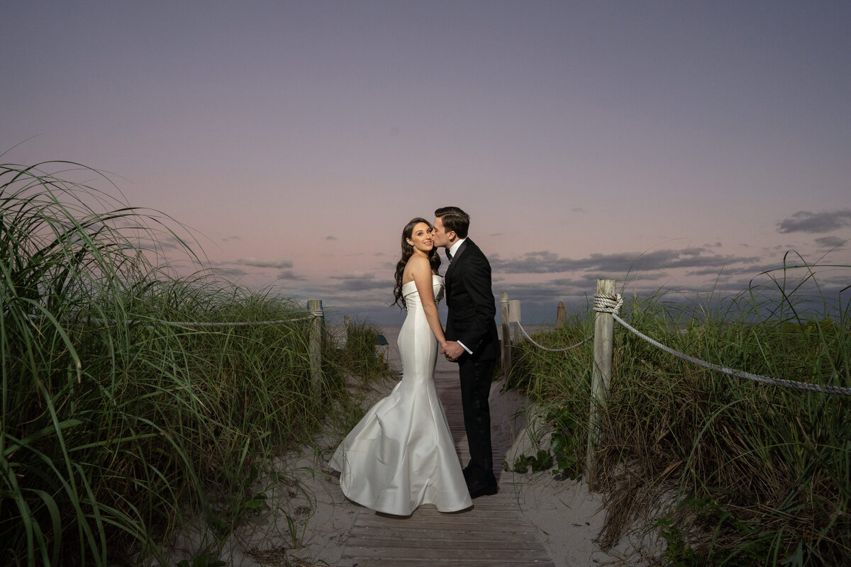 A groom lovingly kisses his bride as they stand together on a beach at sunset, captured by Rodrigo Varela Photography.