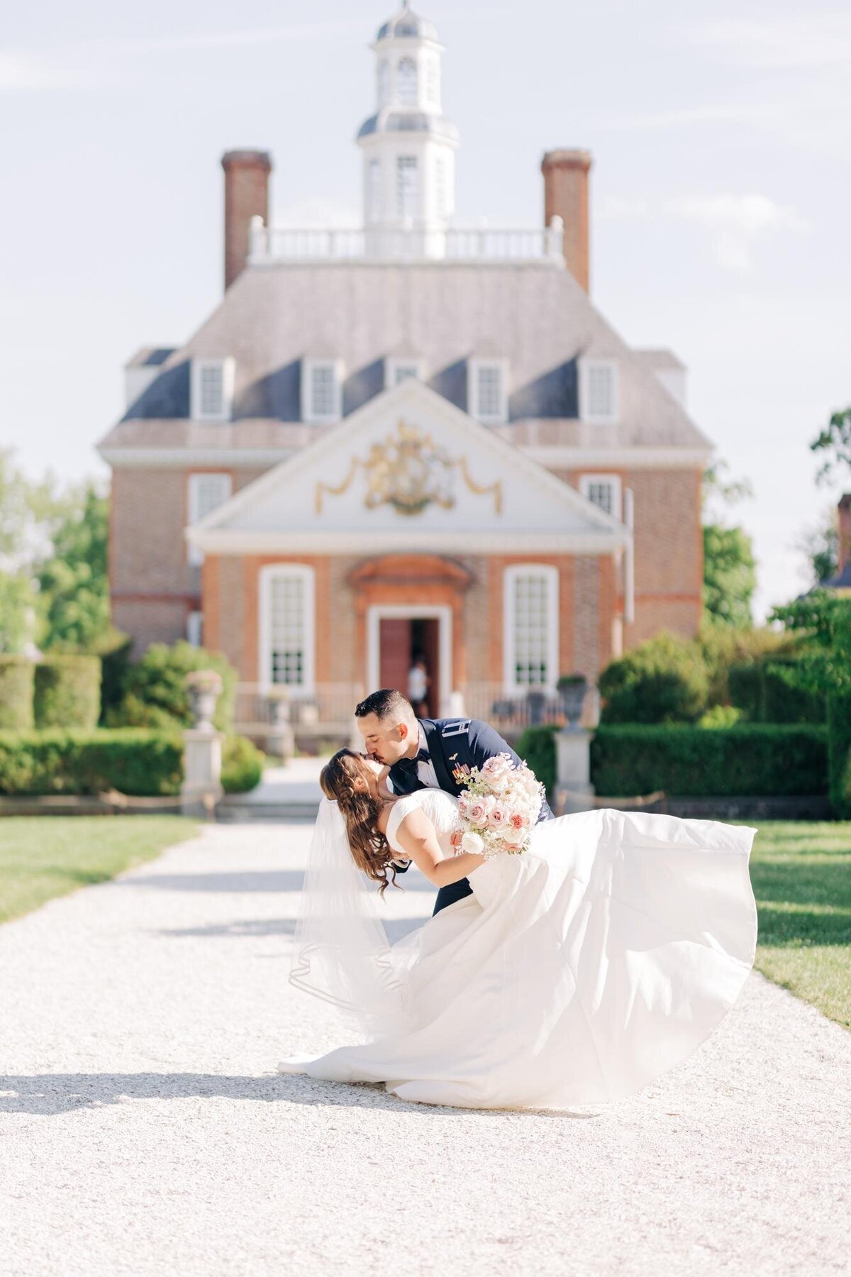 A couple embracing and kissing, with the groom lifting the bride, in front of an elegant manor house on a sunny day.