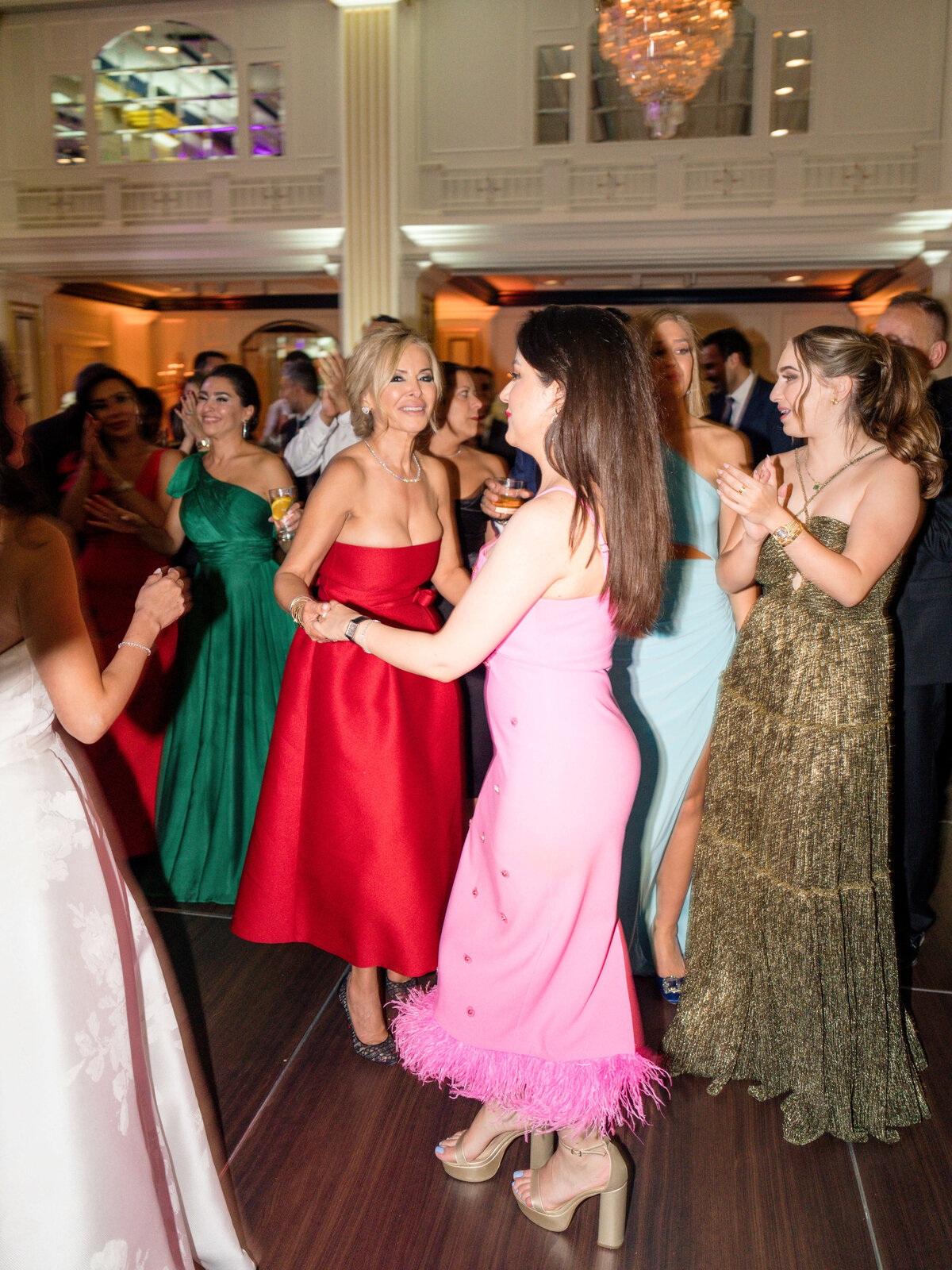 A group of elegantly dressed people dancing at an indoor event. Two women, one in a red dress and the other in a pink dress with feathers, are in the center. Others around them are wearing vibrant dresses, clapping and enjoying the moment.