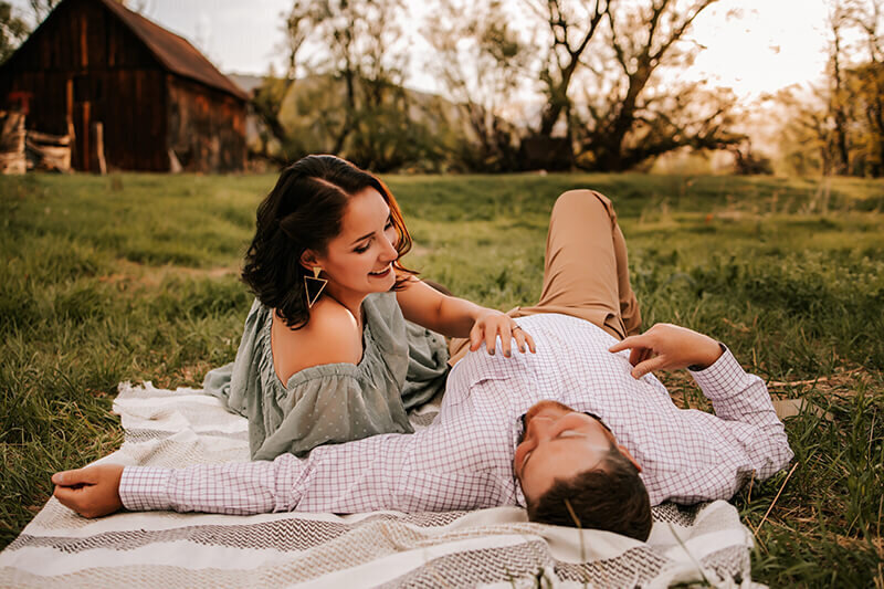 Husband lays on his back while wife leans in next to him in a grassy field in front of a brown barn