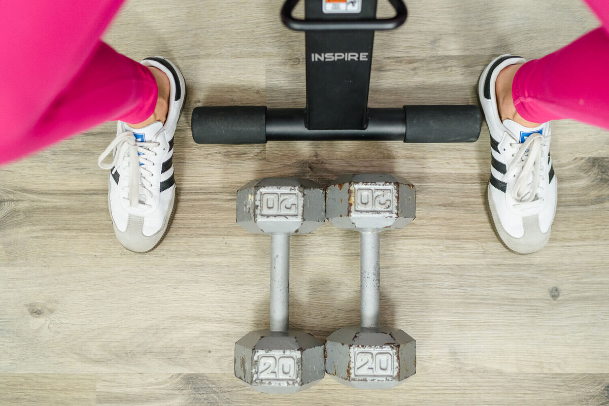 Woman in pink leggings and white Adidas Sambas standing by 20 lb dumbbells and Inspire machine.
