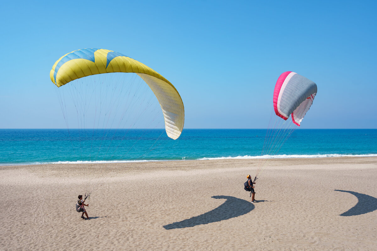 A paraglider soaring above a sandy beach with clear blue skies and the Pacific Ocean in the background, part of a luxury adventure travel experience