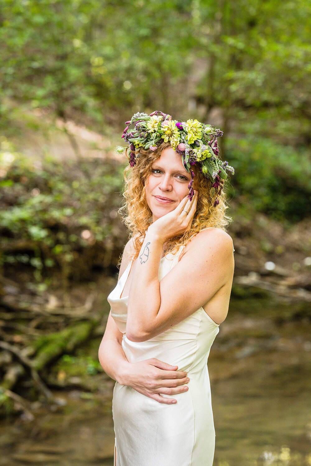 A bride grins while standing in the shallow pond at Fishburn Park for a formal portrait. She is wearing a flower crown on her hand and a simple silk cream dress. She holds onto herself and places her other hand on her face.