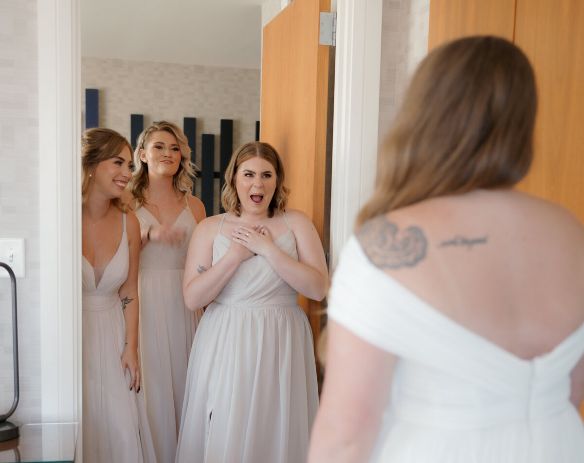 Bride sharing her first look with her bridesmaids, who are smiling and cheering as they see her wedding gown for the first time in an elegant room.