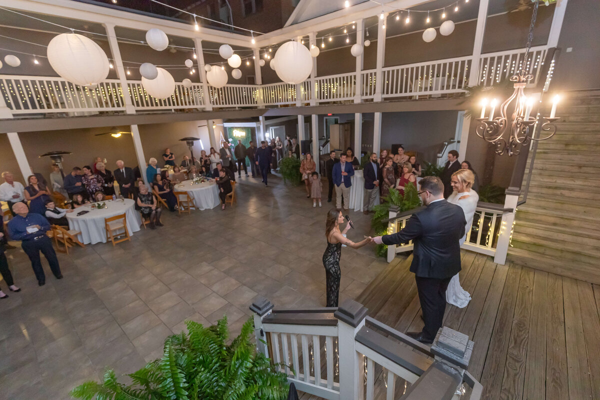 A bride and groom stand on stairs addressing guests at an outdoor wedding reception decorated with string lights and white paper lanterns.