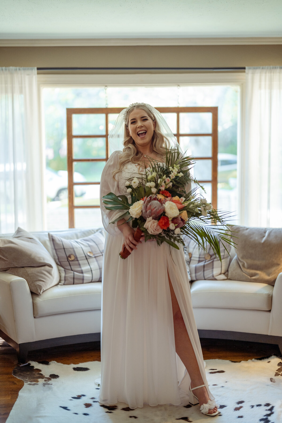 Joyful bride smiling brightly as she holds her bouquet on her wedding day in Fort Worth. Dressed in a flowing white vintage gown with a thigh-high slit, she stands in a beautifully lit room, radiating happiness and excitement for her special day.