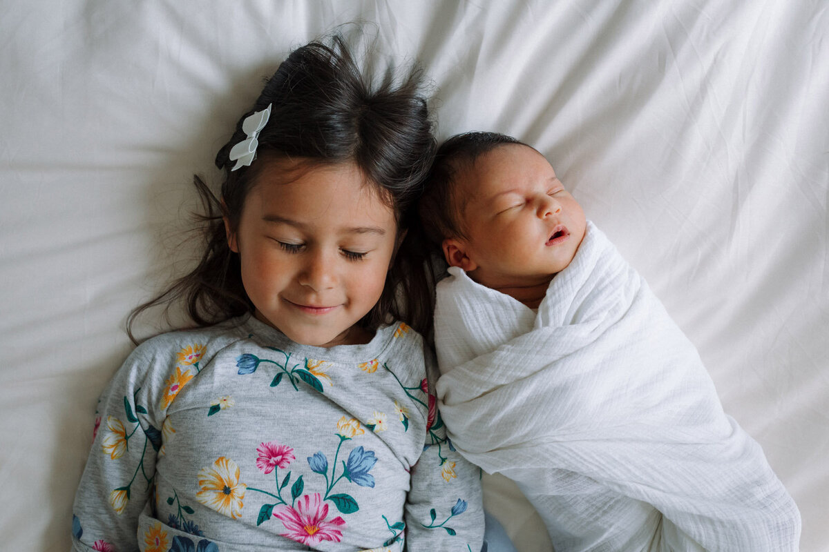 sister in flowered dress with eyes closed laying next to sleeping newborn sister wrapped in white swaddle