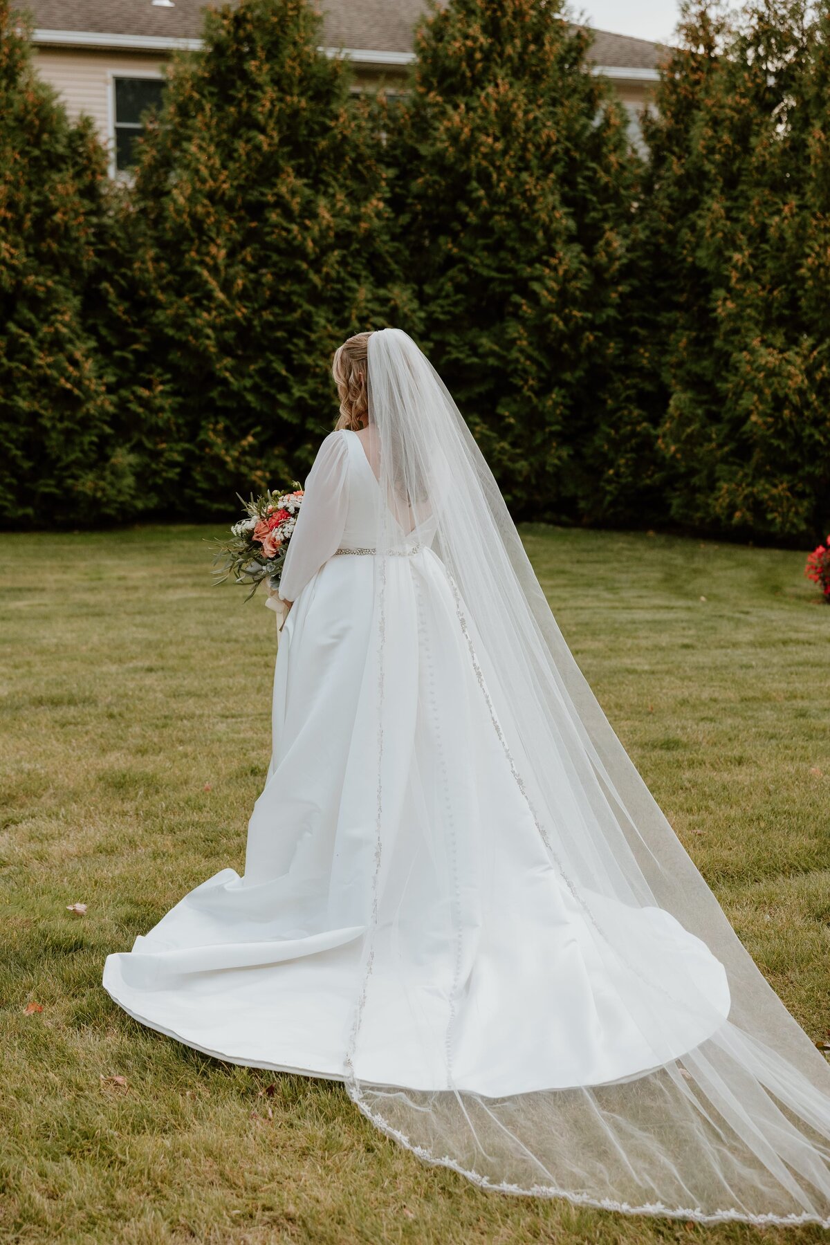 Bride standing on a lawn holding a bouquet, with her long cathedral veil and button-detailed train flowing behind her, photographed from the back against a row of tall evergreens.