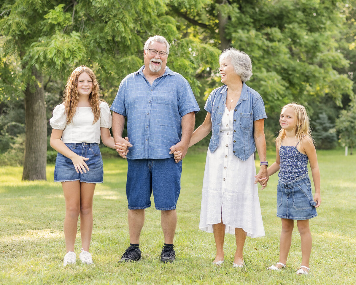 Grandparents and granddaughters standing next to each other, holding hands, in grass in front of a tree line, smiling at each other and the camera.  Older daughter is wearing blue jean shorts, a white short sleeve shirt, and has long brown hair.  Grandpa is wearing a light blue and white stripped short sleeve button up shirt, blue jean shorts, and has white short hair and goatee.  Grandma is wearing a white button up dress with a jean jacket over it, with short white hair.  Youngest daughter has a dark blue and white tank top style shirt with a blue jean skirt, and has medium length blonde hair.