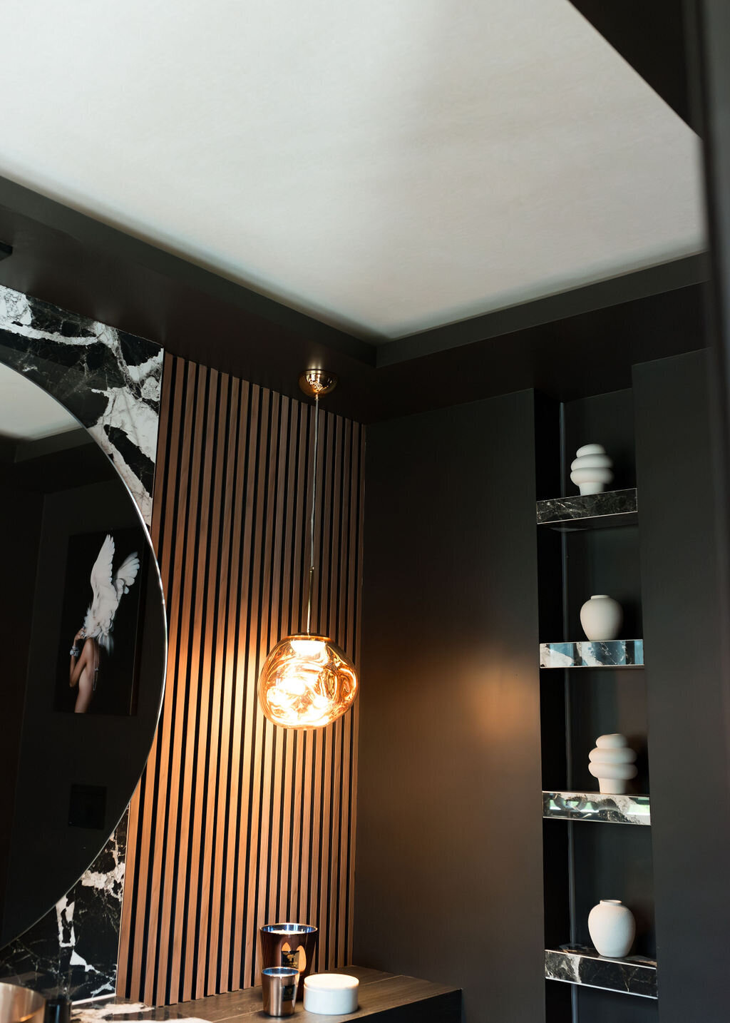 A modern bathroom with dark walls, a large circular mirror, and a glowing pendant light. The wall features vertical wooden slats and marble accents.There are built-in mirrored shelves with decorative white vases on the right side.