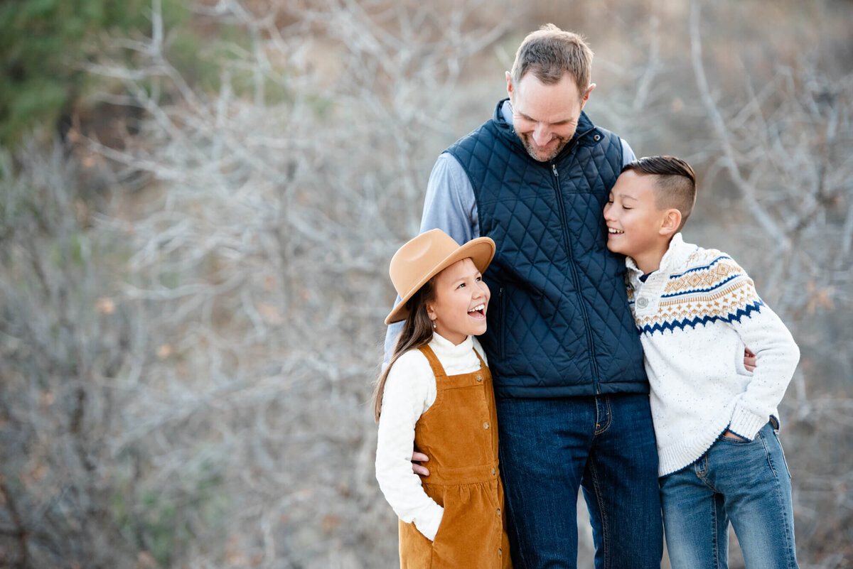 A father stands in a blue vest hugging his young son and daughter as they all laugh