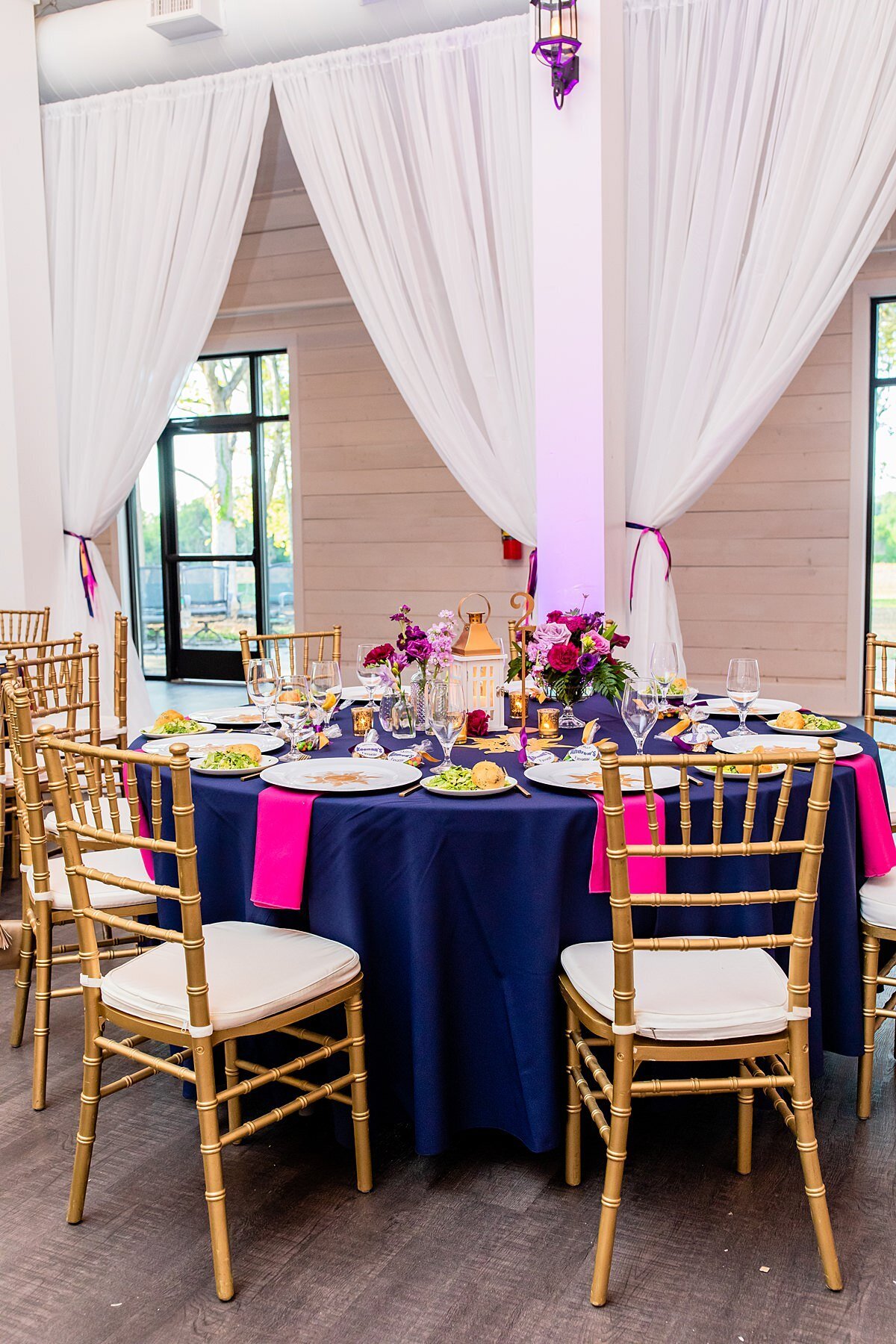 Disney's Tangled wedding table set with a navy linen, hot pink napkins, chargers with gold suns and gold lanterns with pink, purple, magenta flowers and ivory drapery in the background. The table is set with gold chivari chairs with white cushions.