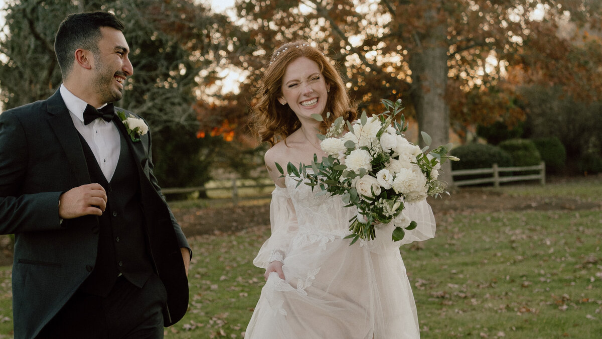 A joyful bride and groom run hand in hand through a park during their wedding day. The bride, holding a lush bouquet of white flowers and greenery, beams with happiness in her off-the-shoulder lace gown, while the groom, dressed in a sharp black tuxedo with a white boutonniere, smiles at her. The autumn foliage and wooden fence in the background add a romantic and picturesque setting.