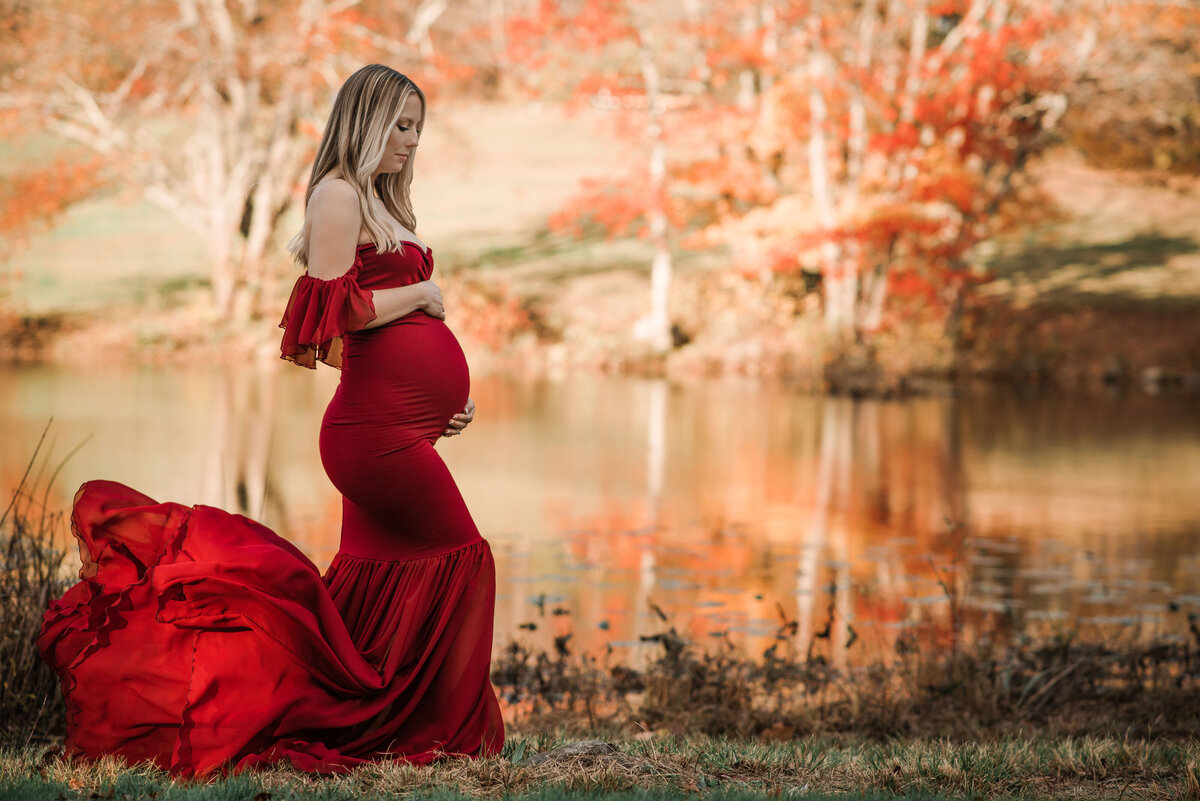 Pregnant woman posing for maternity portrait outdoors in fall leaves wearing a red satin flowing maternity gown holding baby bump and looking down