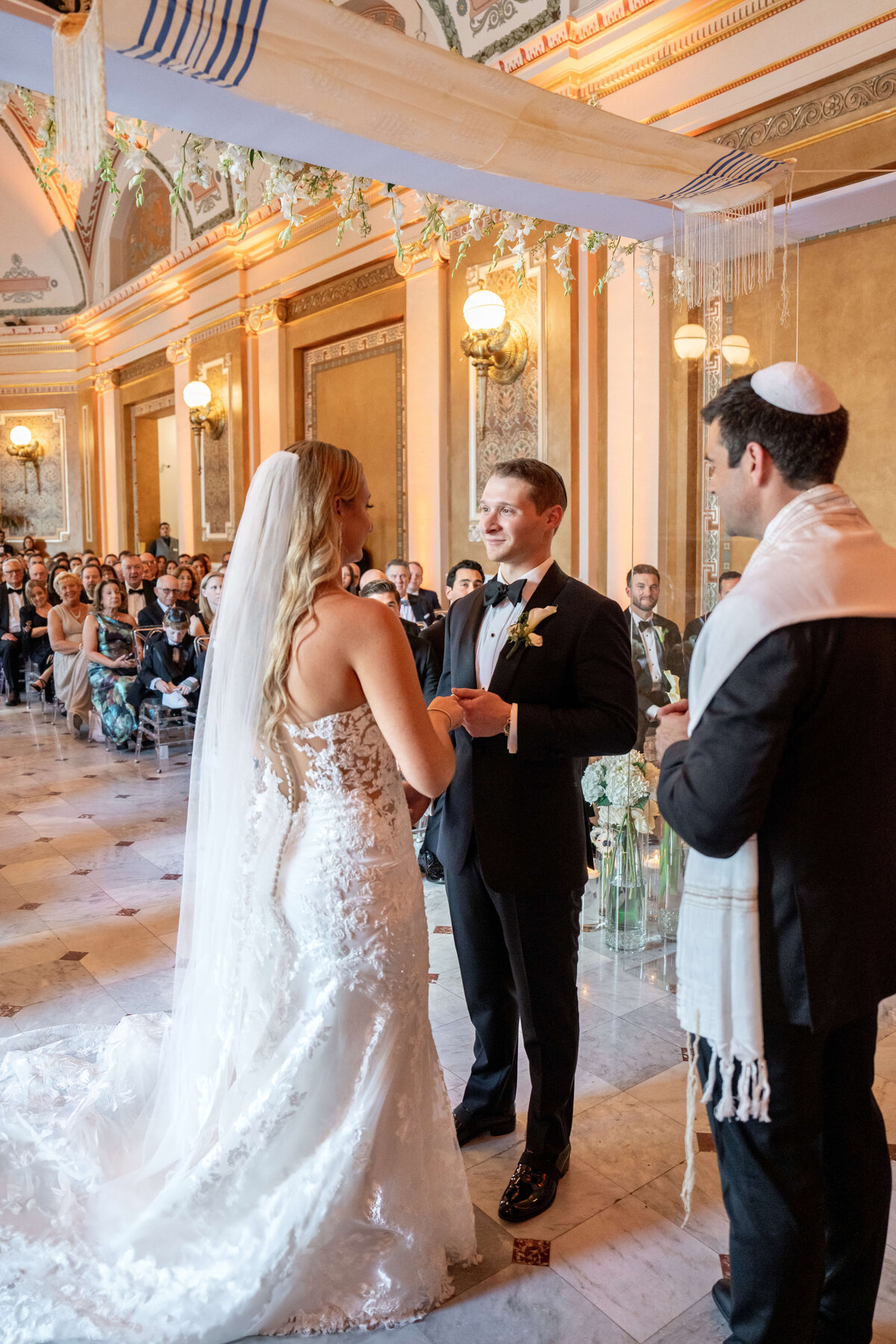 A bride and groom stand under a chuppah during their wedding ceremony in an ornate hall. The bride wears a lace gown and veil, while the groom is in a black suit. A man officiates, wearing a kippah and prayer shawl. Guests are seated in the background.