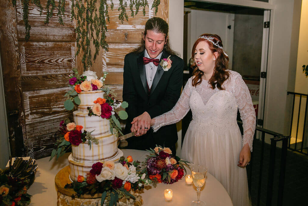 A bride and groom, dressed in wedding attire, smile as they cut and serve each other a piece of a three-tiered white wedding cake during their reception in a softly lit room.