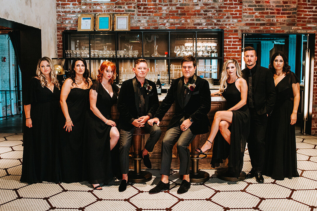 Two grooms wearing black tuxedos pose indoors with their wedding party in front of a brick wall..