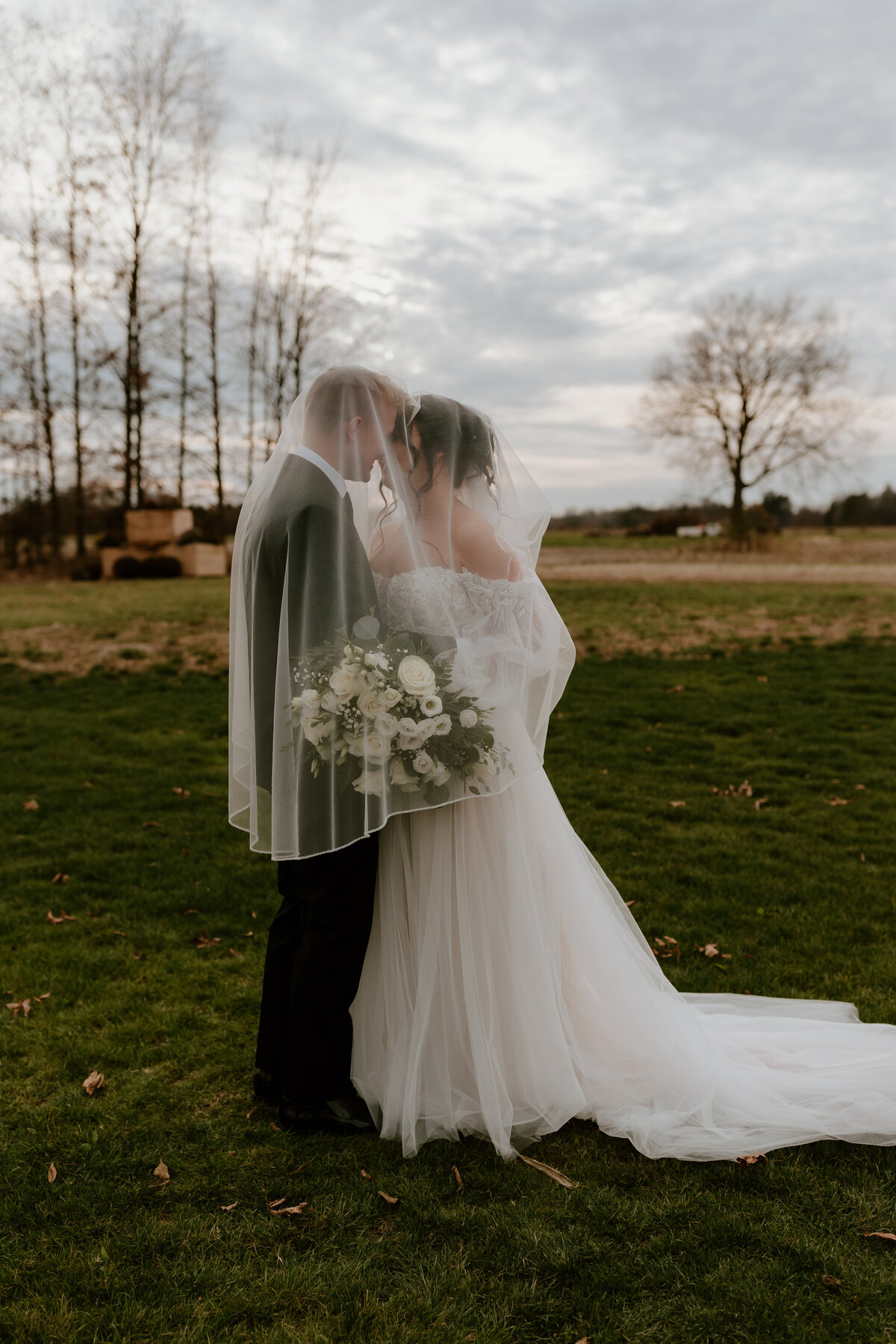 A bride and groom share a romantic kiss, surrounded by tall trees and soft grass, with the bride holding a lush bouquet.