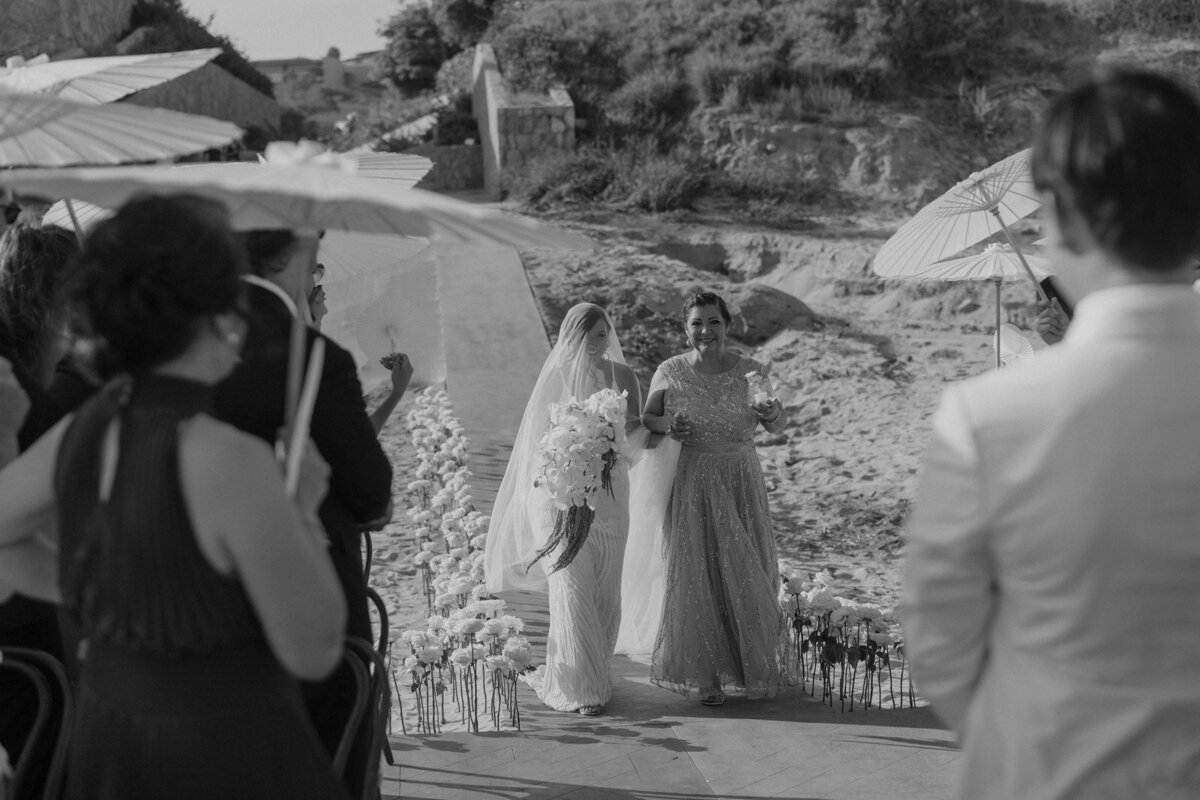 Bride walks down the aisle at a beach wedding in Mexico wearing all white with white flowers, a long veil and her mom at her side. Beach umbrellas are nearby.
