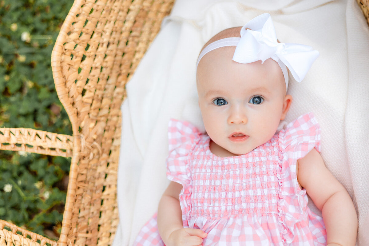 4 month old baby girl wearing a pink and white dress laying in a moses basket looking at the camera