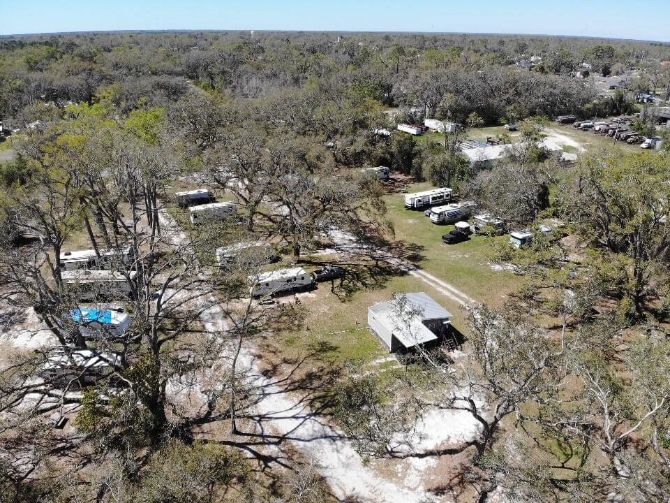 Aerial view of Mayo Florida campgrounds with several RVs and scattered trees. Dirt roads meander through the site near the Suwannee River Rendezvous camping area, where a pavilion is visible. Sparse vegetation suggests a rural Lafayette County setting under a clear sky.