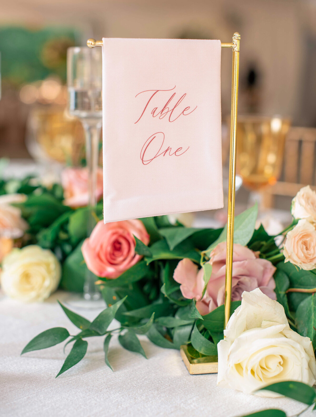 A small table flag reads Table One in elegant script, surrounded by pink and white roses with green leaves. Gold-rimmed wine glasses are visible in the background, adding a touch of elegance to the setting.