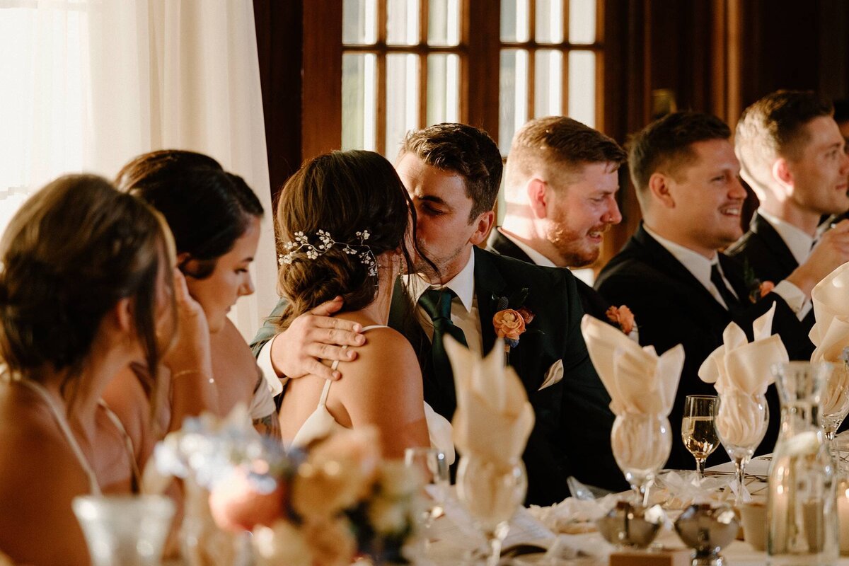 a bride and groom kissing amongst their wedding party while seated at the head table on their wedding day