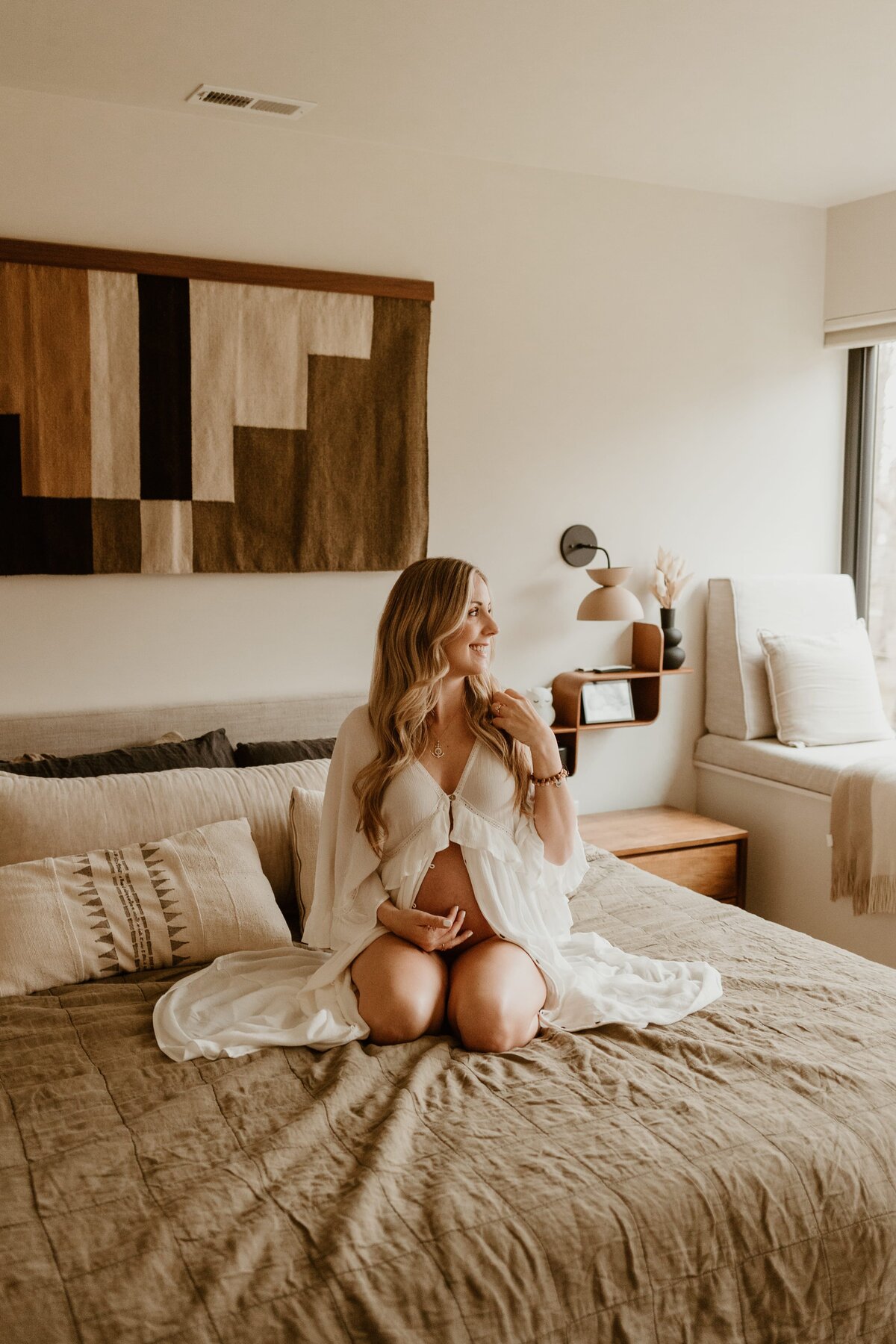 A cheerful pregnant woman sitting cross-legged on a bed, holding her belly, with a neutral-toned, cozy bedroom decor in the background.