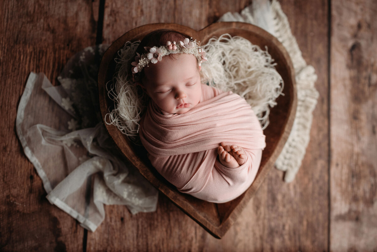 Newborn girl in dusty pink swaddle with feet popping out, hand tucked under chin, wearing floral headpiece and laying in a wooden heart shaped bowl set on dark brown hardwood flooring