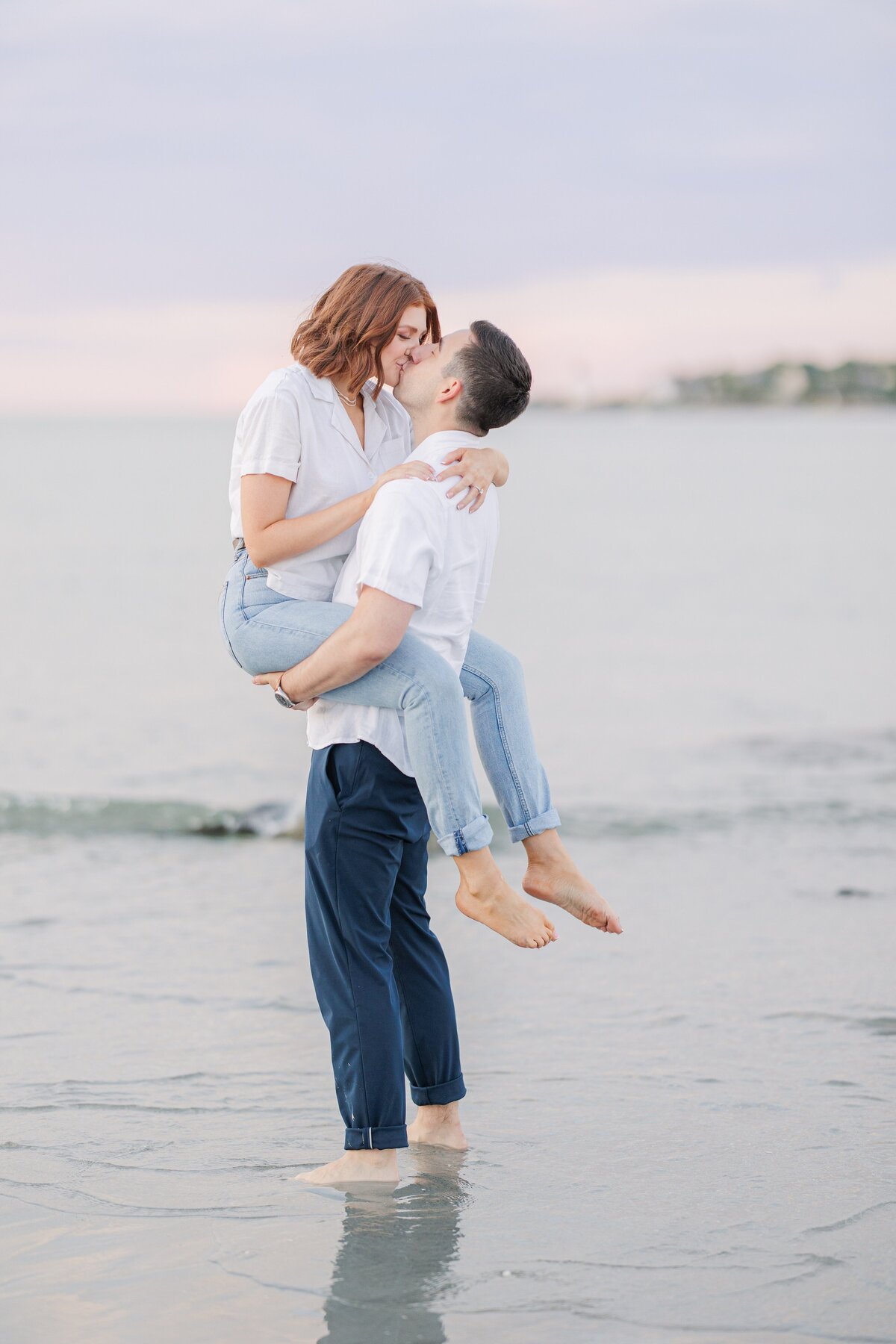 A man picking up a woman and sharing a kiss as they stand in the waves at Wingaersheek Beach during their sunset engagement photos.