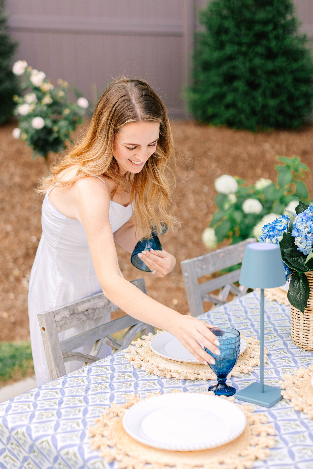 A woman in a white sundress sets blue glassware on an outdoor dining table decorated with a patterned tablecloth, woven placemats, and a hydrangea centerpiece. She smiles while arranging the table, with blooming flowers and greenery in the background.