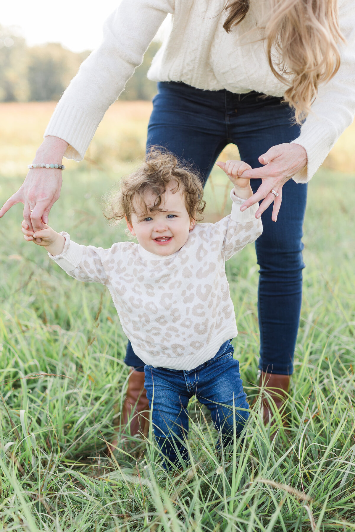 A northern virginia family photographer photo of a mother helping her daughter walk through the grass while smiling at the camera