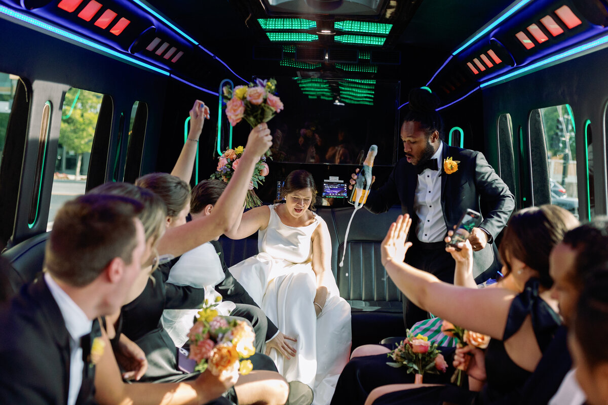 Bride, groom, and wedding party cheering with drinks on a luxury party bus after Washington DC wedding ceremony.